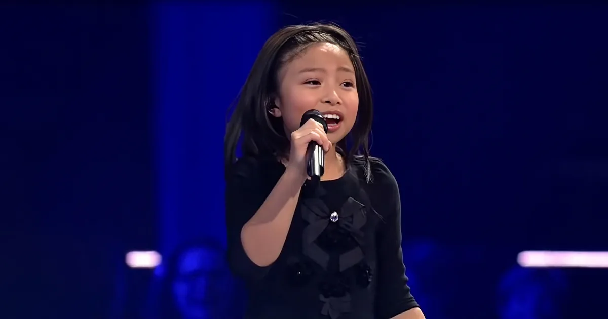 A young girl in a black dress sings into a microphone on stage with a dark blue background.