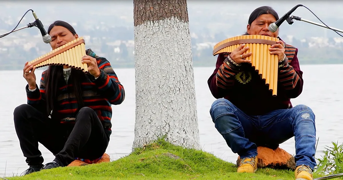 Two individuals play panpipes by a lake, one in striped sweater, the other in jeans, both focused on their music.