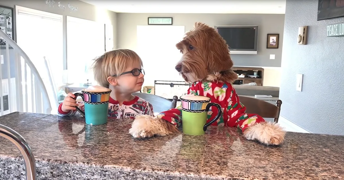 A child and a dog in pajamas sit at a kitchen counter, each holding a mug, looking at each other.