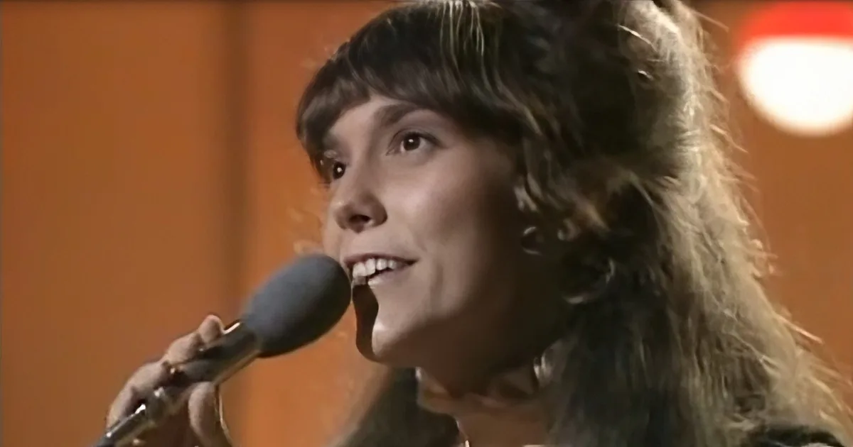 A person with long, wavy hair sings into a microphone against an orange background.