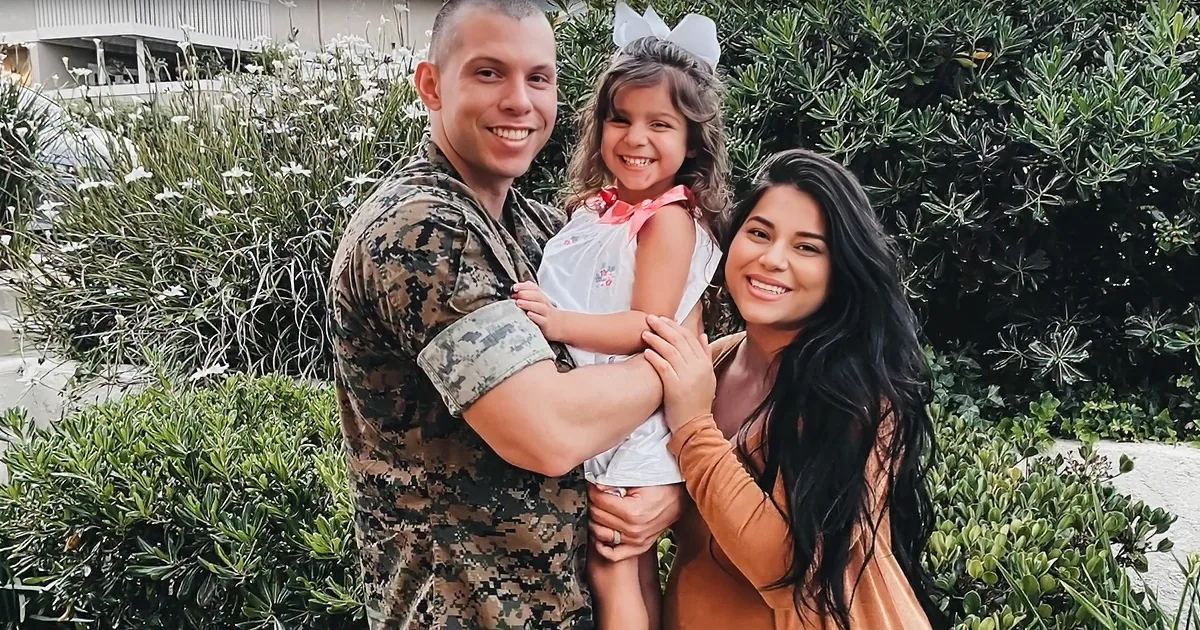 A soldier in camouflage holds a smiling child, with a woman in a brown top beside them, all outdoors with greenery.