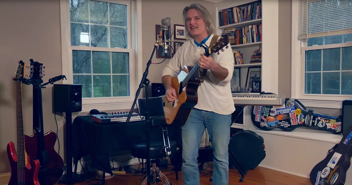 A man plays guitar in a room with musical equipment, books, and windows.