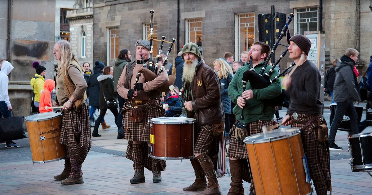 A group of men in kilts and traditional attire play bagpipes and drums on a city street.