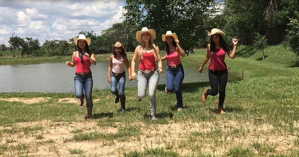 Five women in cowboy hats and jeans run together on a grassy field near a lake.