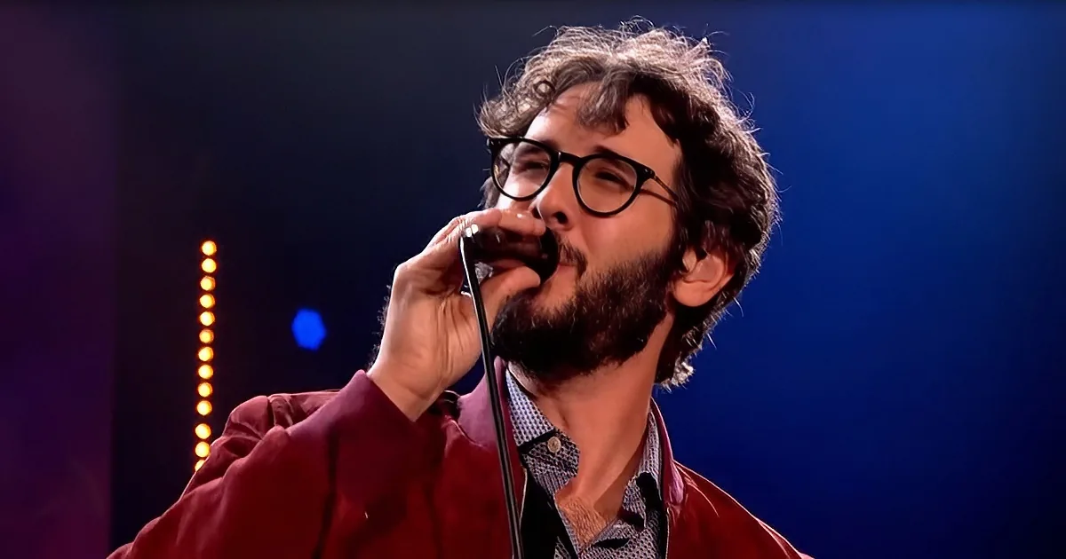 A man with glasses and a beard, wearing a red jacket, holds a microphone close to his mouth.