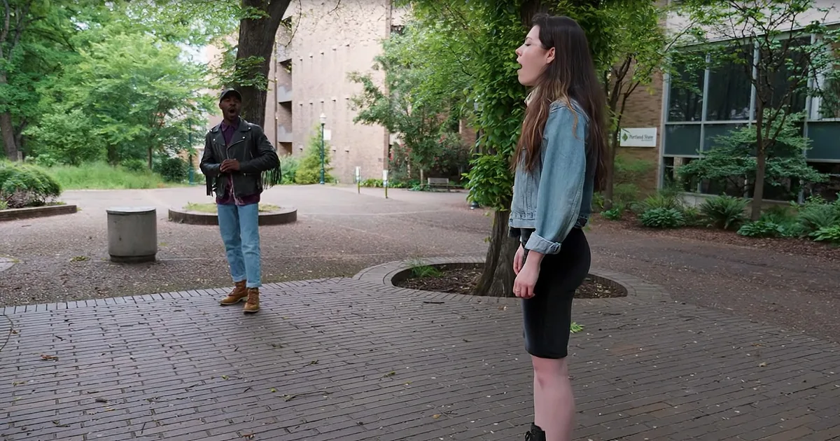 A woman in a denim jacket and black skirt stands on a brick path, facing a man in a leather jacket and jeans who is speaking.