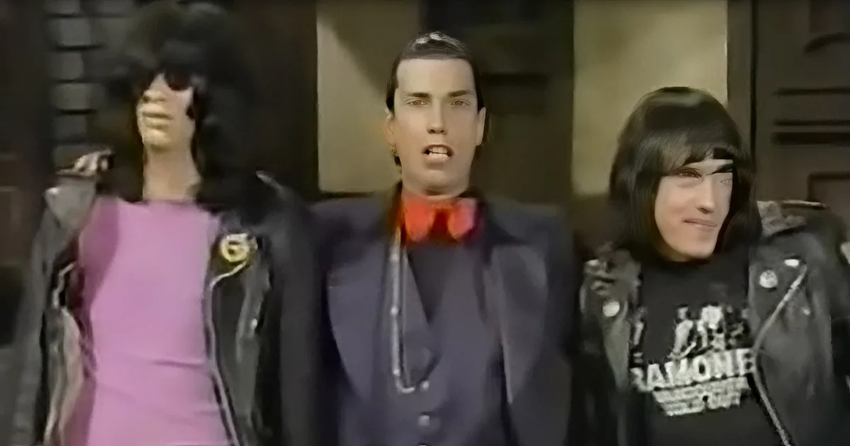 A man in a suit and bow tie stands between two individuals in leather jackets, one in a pink dress and the other in a...