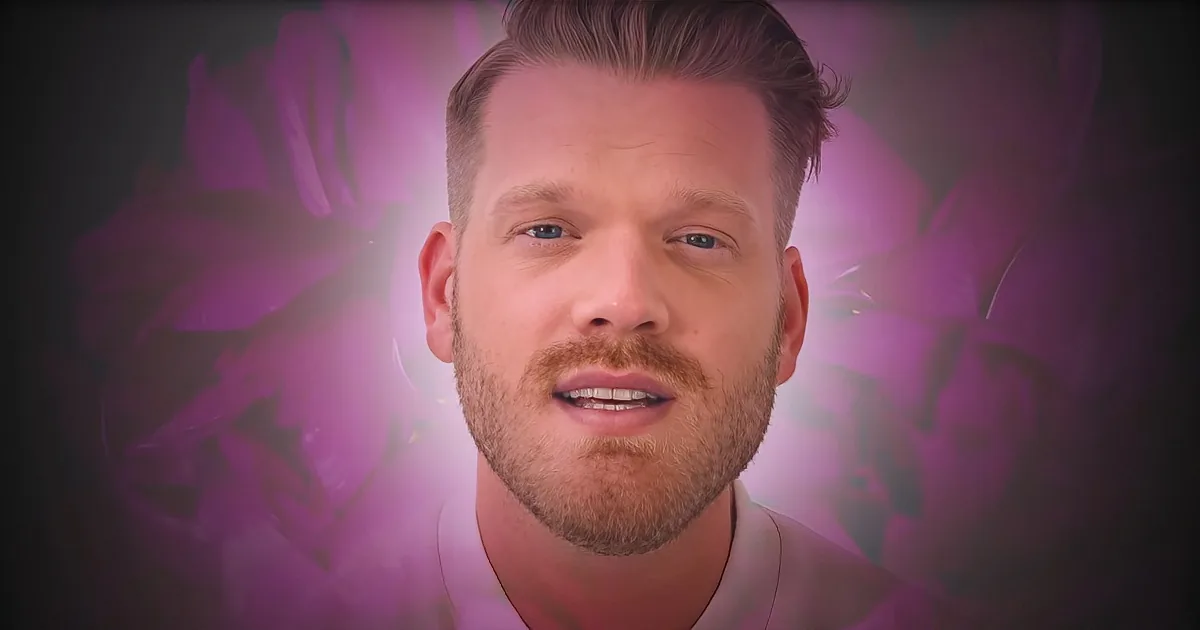 A man with short hair and a beard, wearing a white shirt, is speaking in front of a purple floral background.