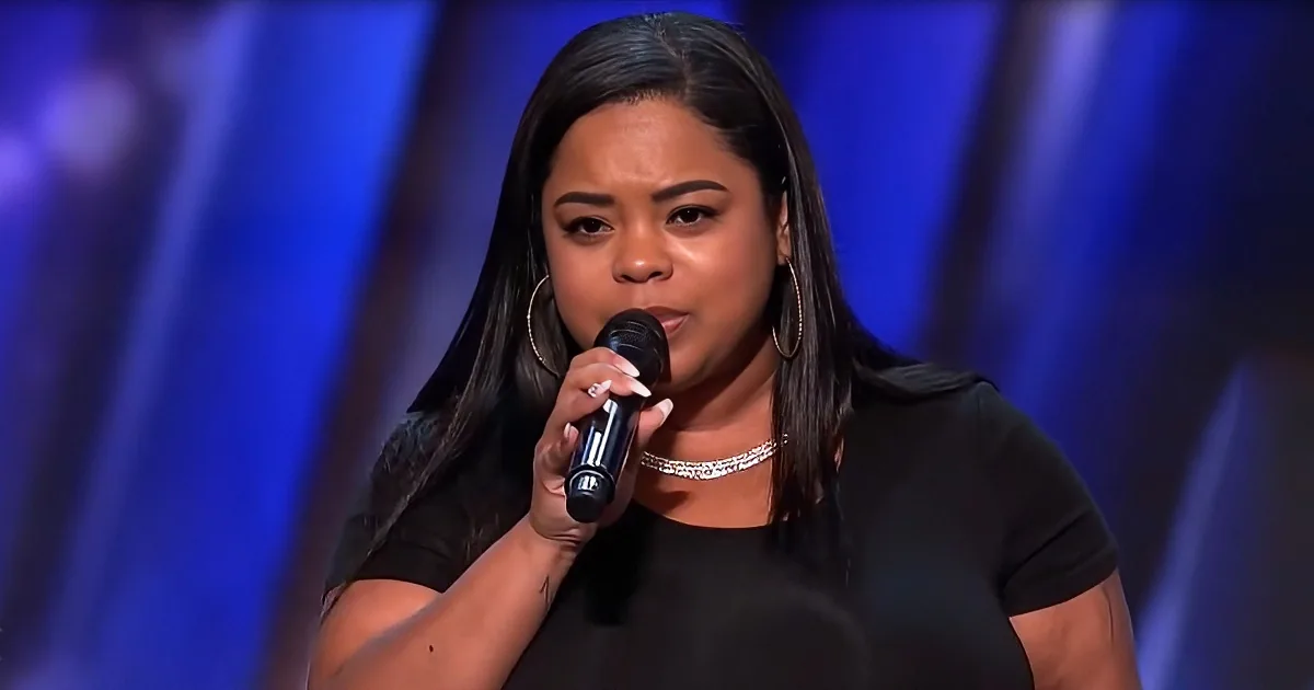 A woman with long dark hair, wearing a black top and a choker necklace, sings into a microphone on a stage with blue...