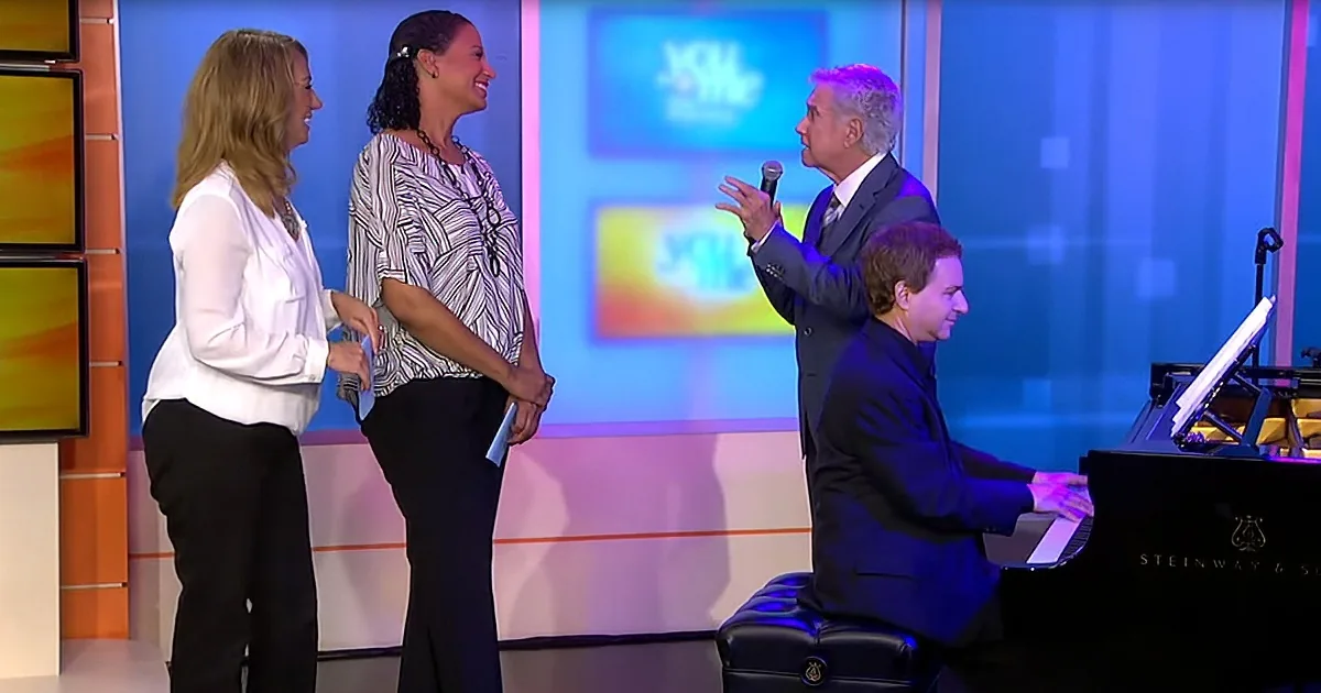 A man in a suit speaks into a microphone while a woman shakes hands with another woman, both smiling.