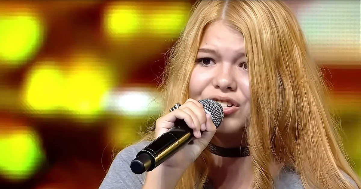 A person with long blonde hair sings into a microphone, wearing a choker, against a blurred colorful background.