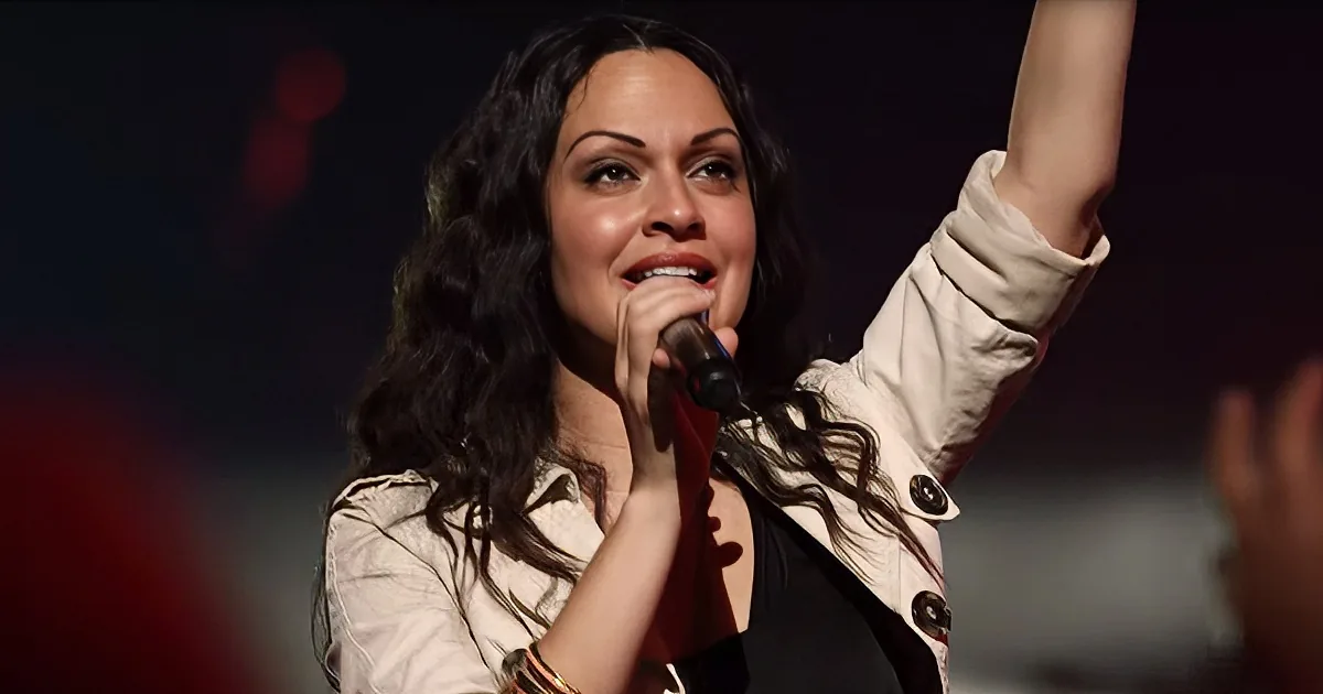 A woman with long dark hair sings into a microphone, raising one arm in the air.