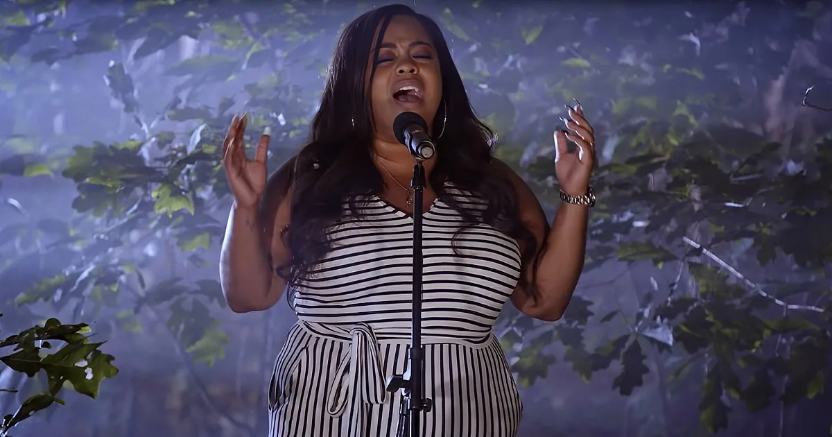 A woman in a striped dress sings passionately into a microphone, surrounded by a misty, leafy background.