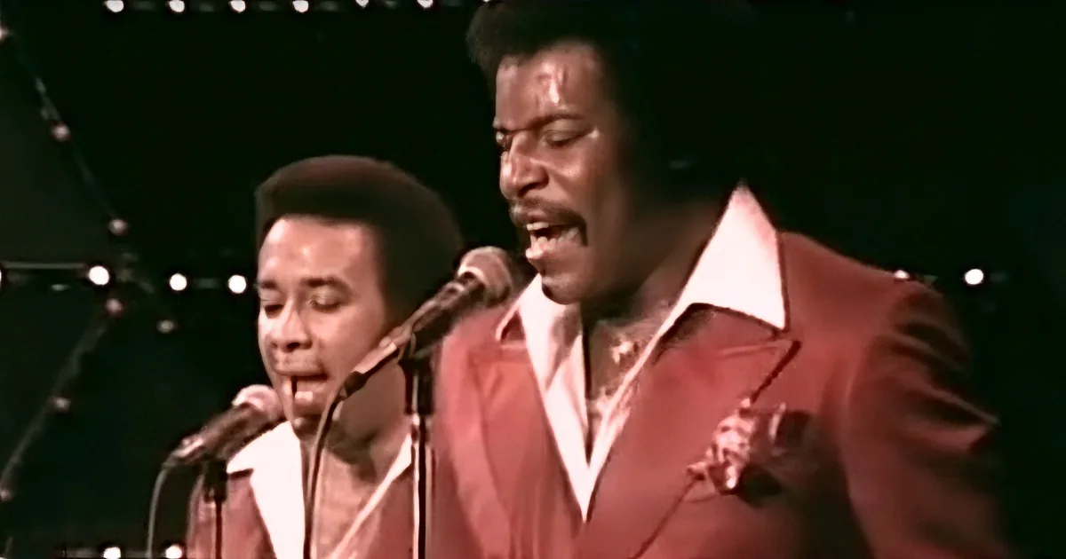 A man in a red suit sings passionately into a microphone on stage.
