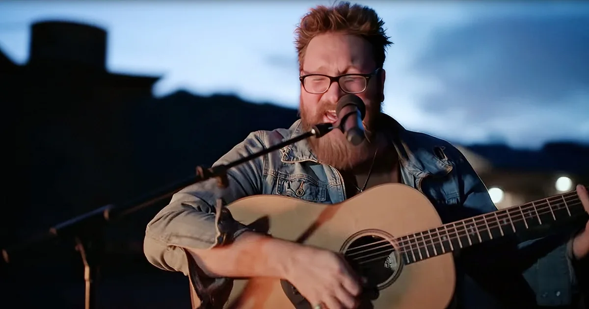A man with glasses and a beard plays an acoustic guitar while singing into a microphone.