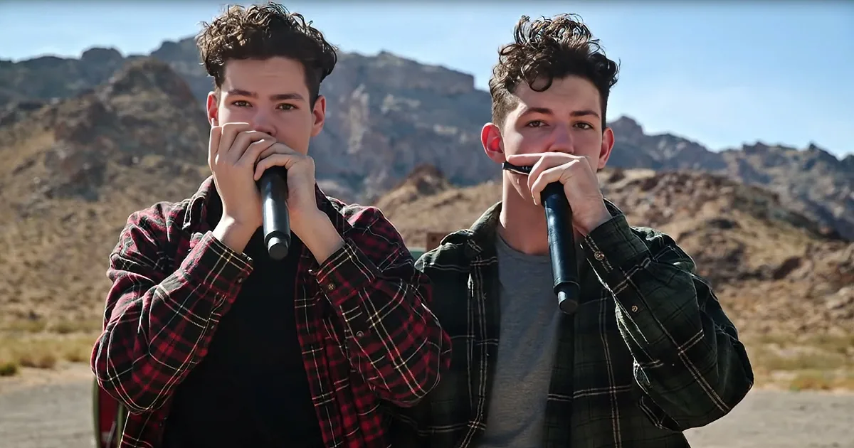 Two individuals in plaid shirts play harmonicas in a desert landscape with rocky hills in the background.
