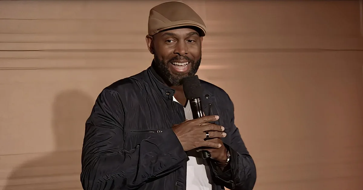 A man wearing a cap and black jacket holds a microphone, smiling, against a beige background.
