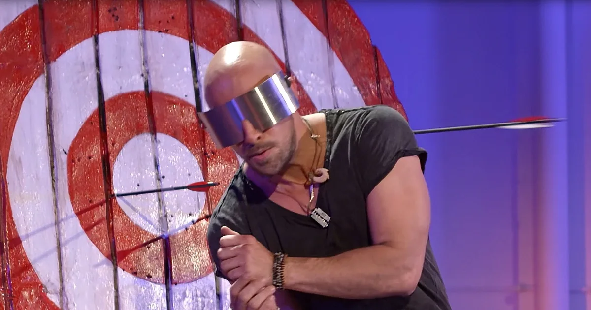 A man with a metallic blindfold stands in front of a target with an arrow hitting it.