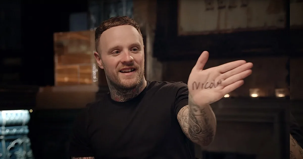 A man with tattoos and a black shirt gestures with his hand, which has "Nicola" written on it.