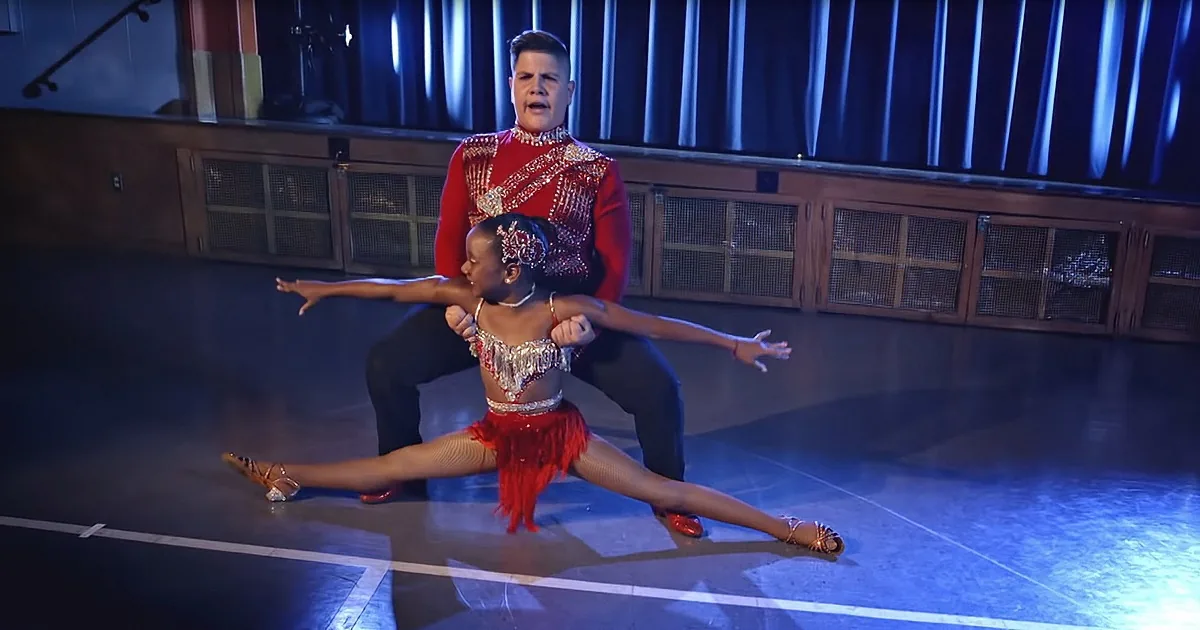 A dancer in red attire performs a split while another dancer supports them in a dance studio.