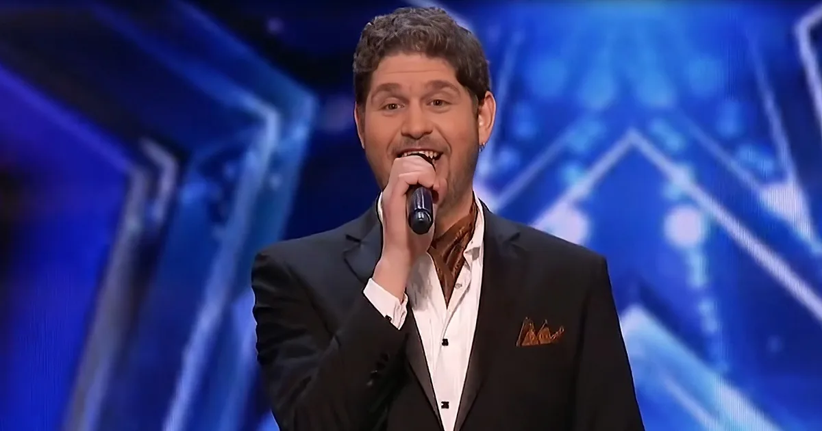 A man in a suit sings into a microphone on a stage with a blue background.