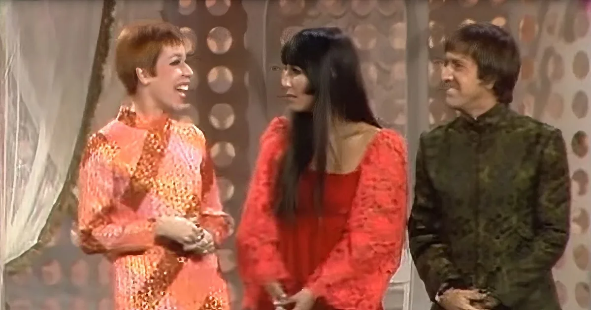 A woman in a red dress stands between two men, one in a patterned orange outfit and the other in a green jacket, in a...