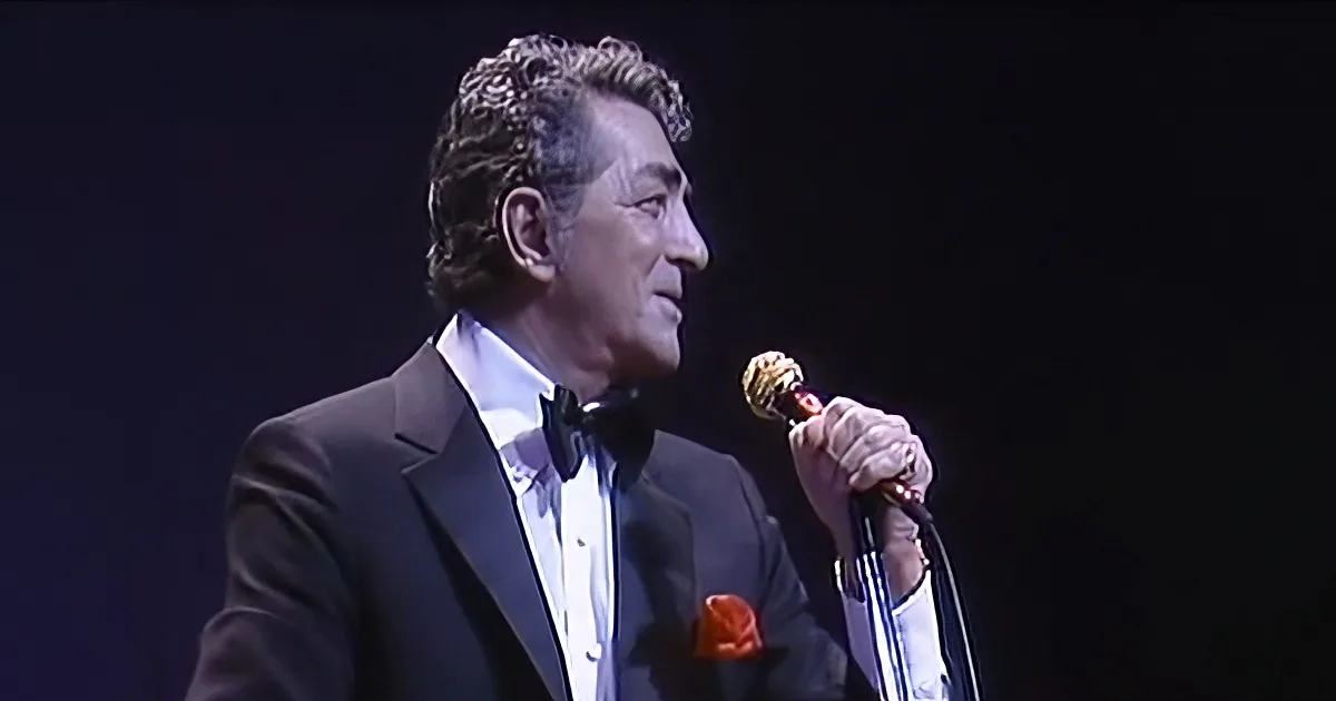 A man in a tuxedo sings into a microphone on stage.