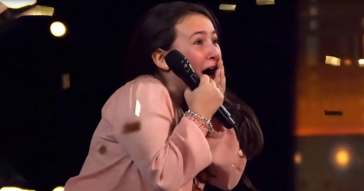 A woman in a pink jacket sings passionately into a microphone on stage.