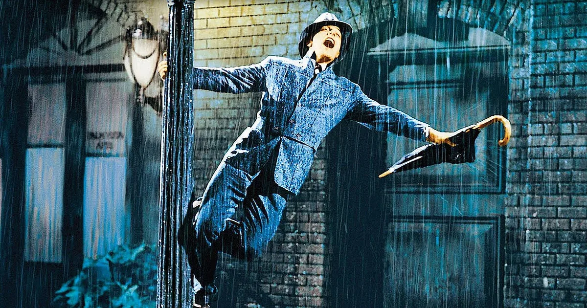 A man in a blue suit dances joyfully in the rain, holding an umbrella, against a brick building backdrop.