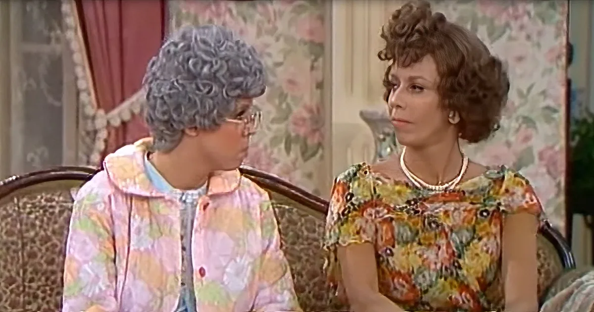 Two women in floral attire sit on a couch, one with glasses, in a room with floral wallpaper and curtains.