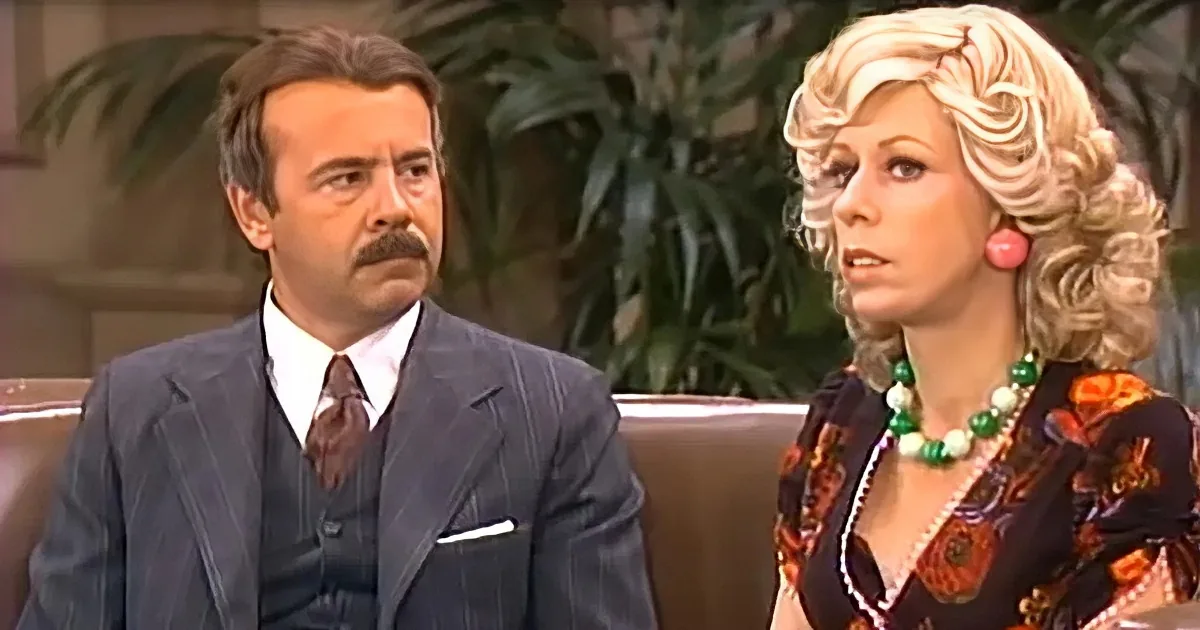 A man in a suit and a woman in a floral dress sit on a couch, both looking off to the side.