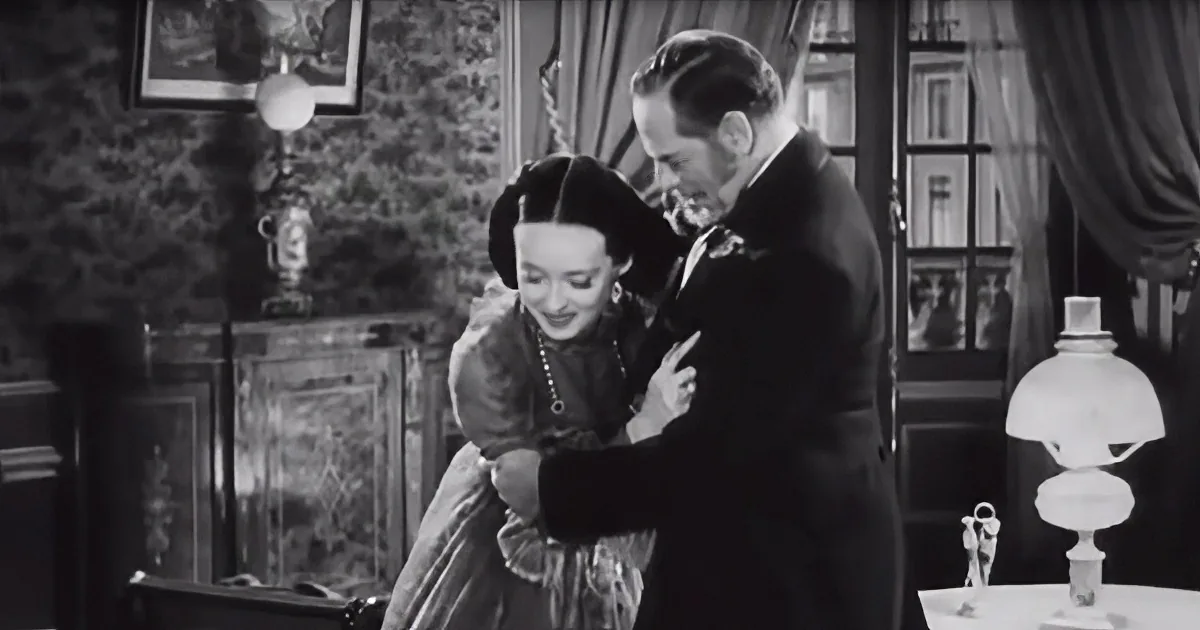 A man in a suit helps a woman in a dress adjust her dress in a vintage room.