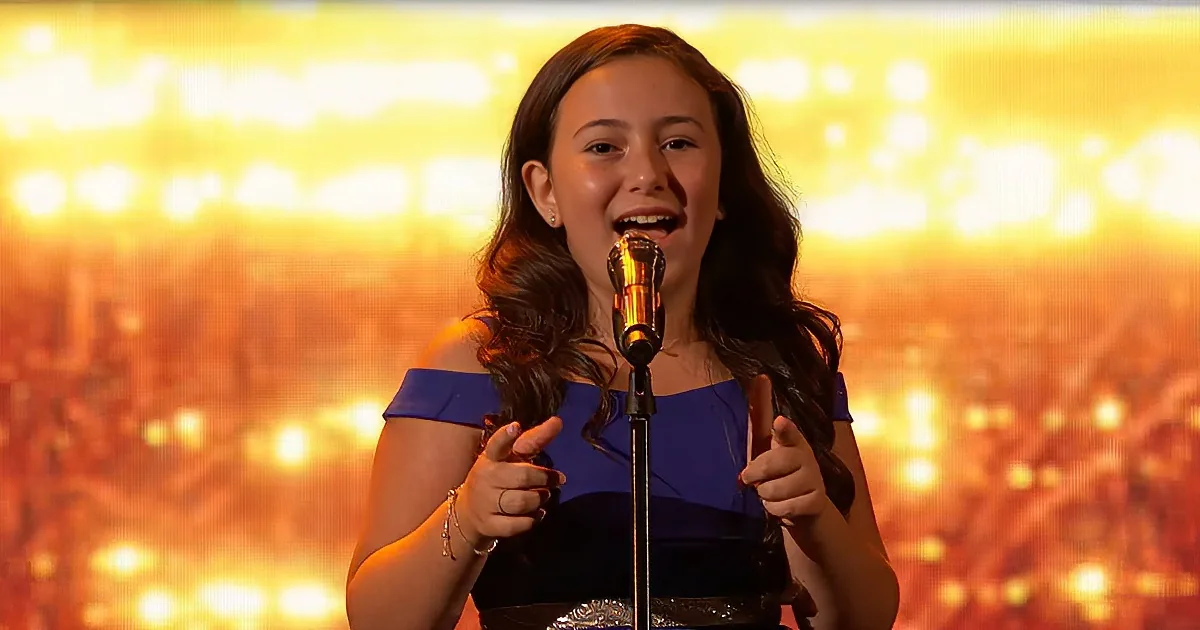 A girl in a blue dress sings into a microphone, smiling and gesturing with her hands.