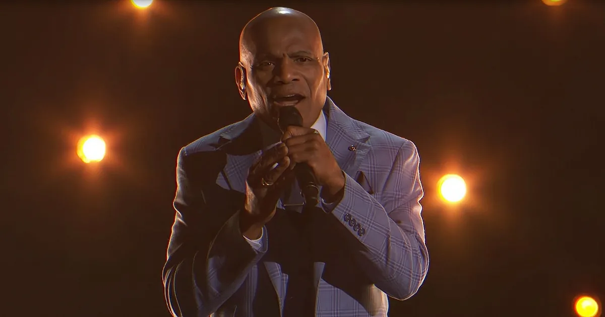 A man in a checkered suit sings into a microphone under warm stage lights.