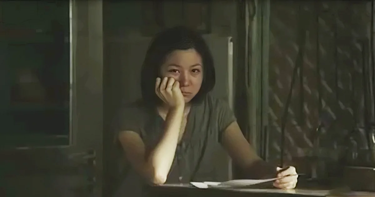 A woman sits at a table, hand on face, appearing contemplative or distressed.