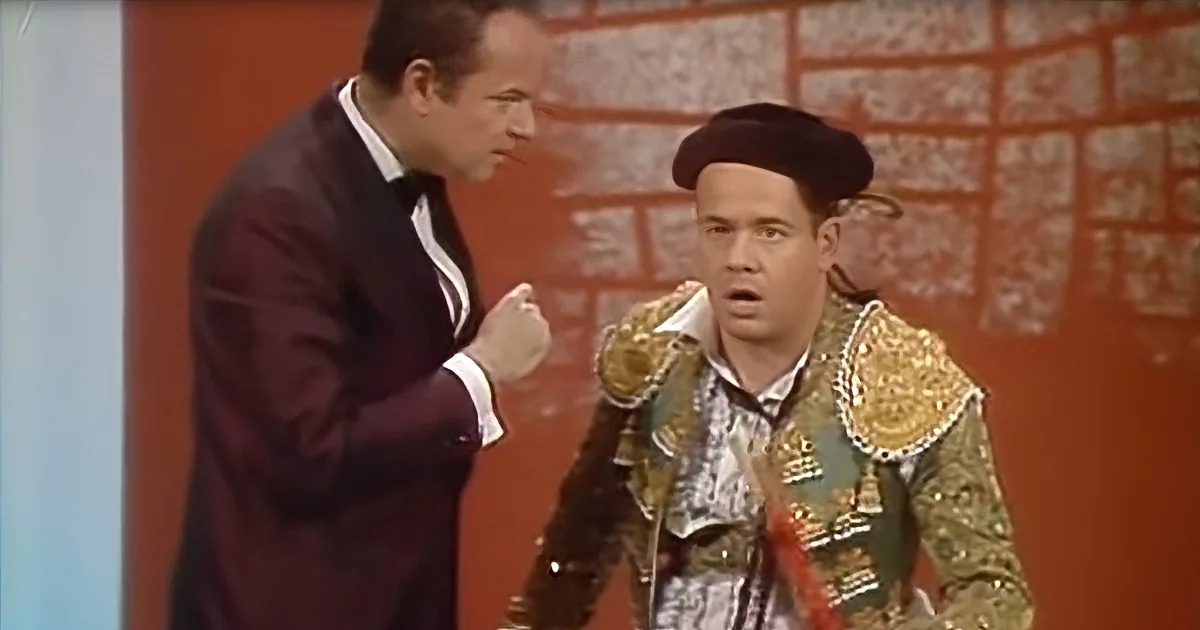 A man in a suit gestures while another in a matador outfit looks surprised.