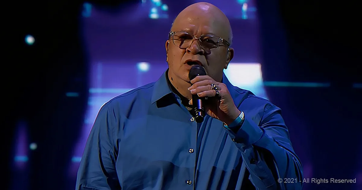 A man in a blue shirt sings into a microphone on stage with purple lighting.