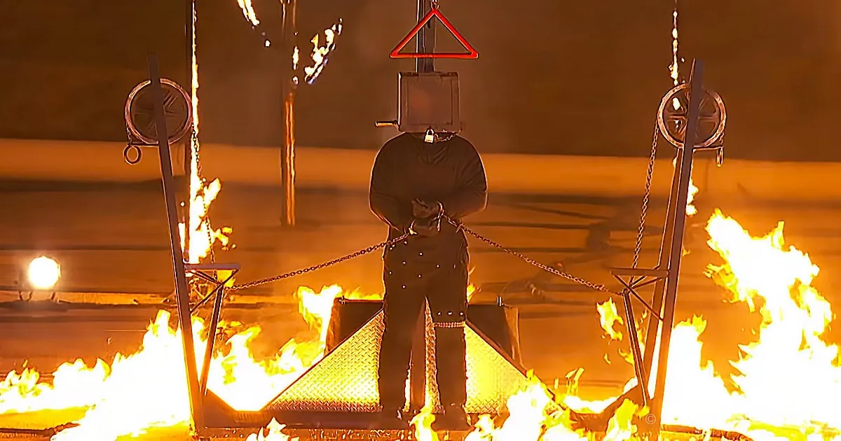 A person in protective gear stands on a platform surrounded by flames, with a red triangular sign above their head.