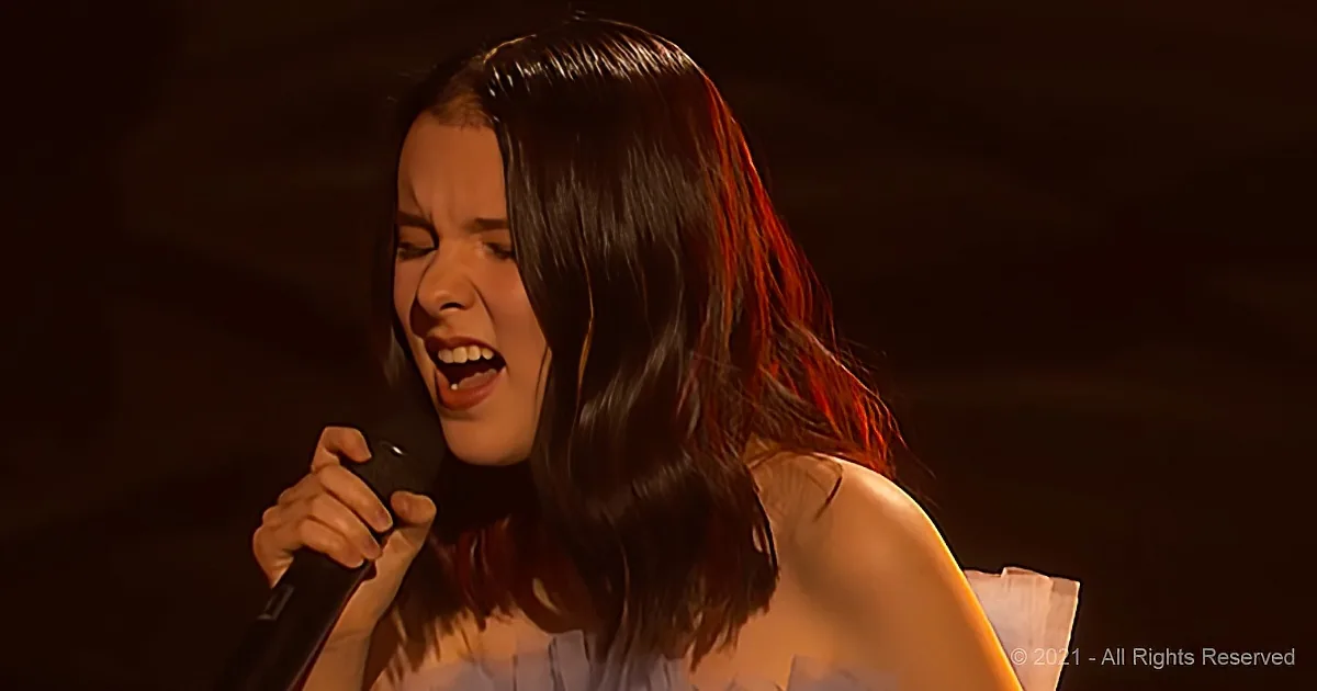 A woman with long dark hair sings passionately into a microphone, illuminated by stage lighting.