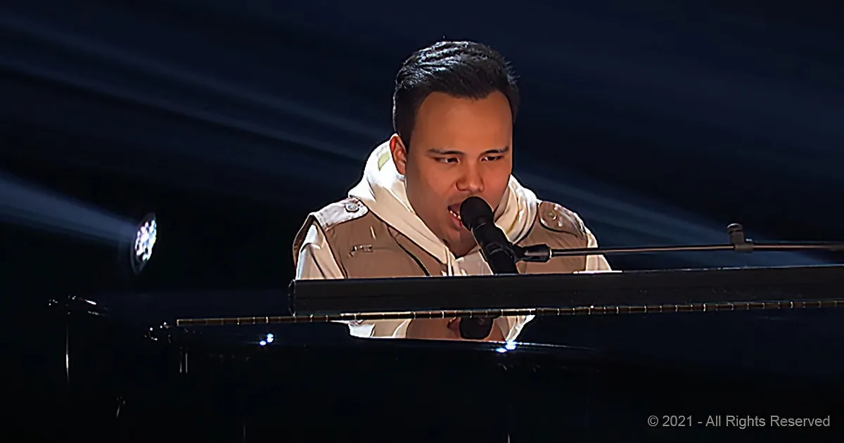 A person in a beige jacket sings into a microphone while playing a piano.