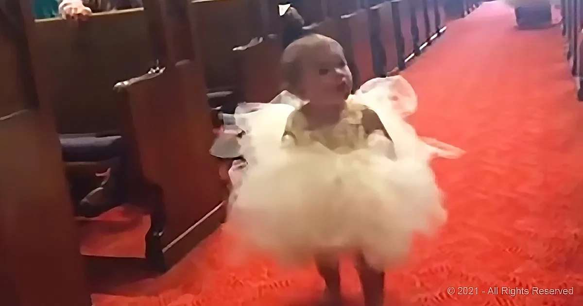 A baby in a white dress sits on a red carpet, surrounded by chairs and people in the background.