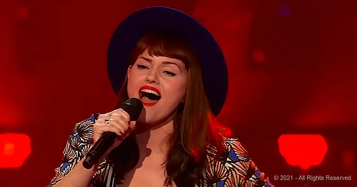 A woman in a hat and patterned top sings into a microphone against a red backdrop.