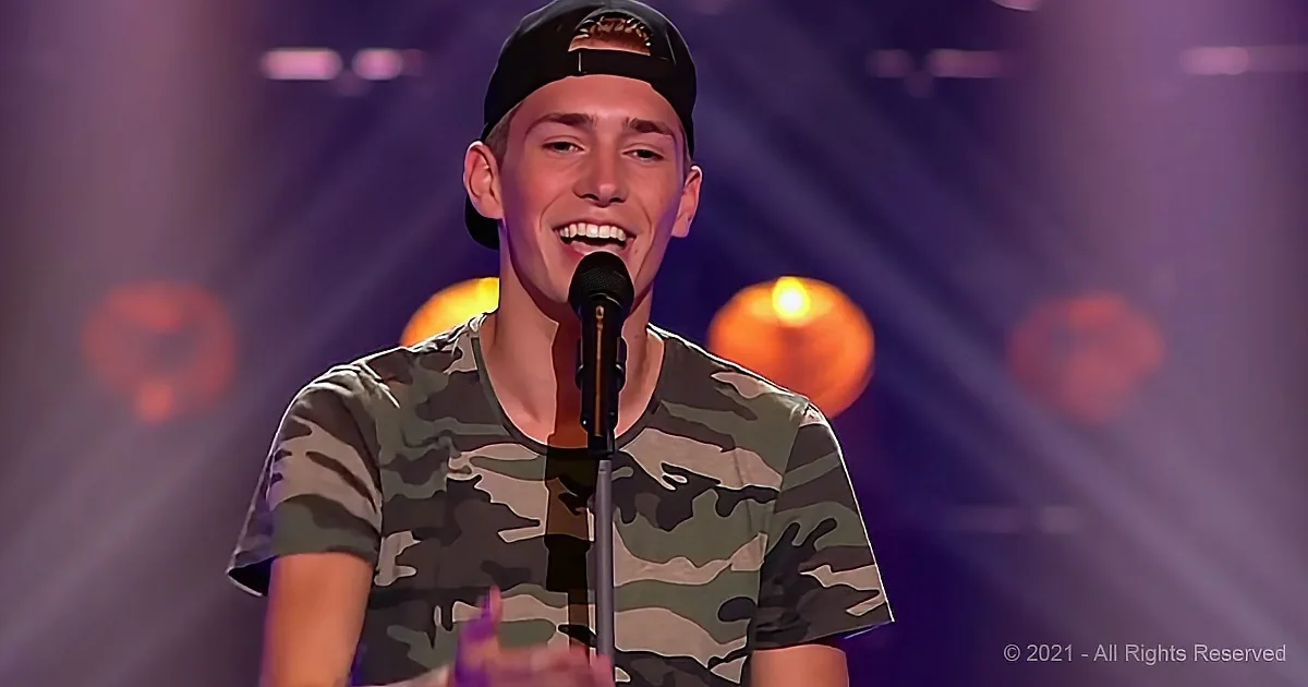 A young man in a camouflage shirt and cap smiles while holding a microphone on stage.