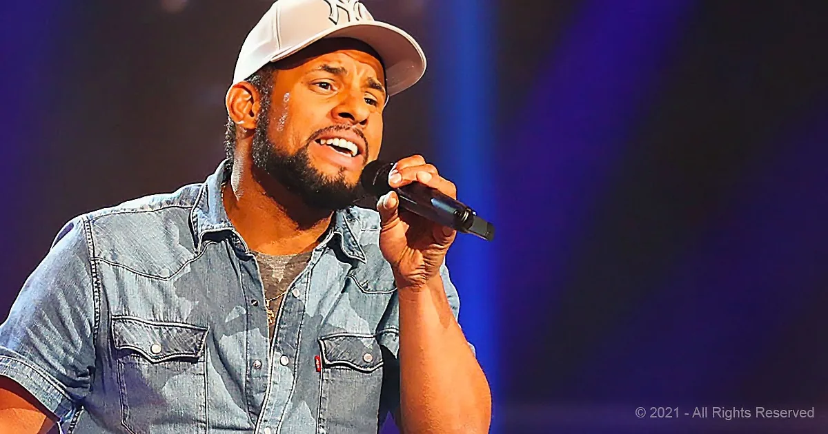 A man in a denim shirt and Yankees cap sings into a microphone on stage.