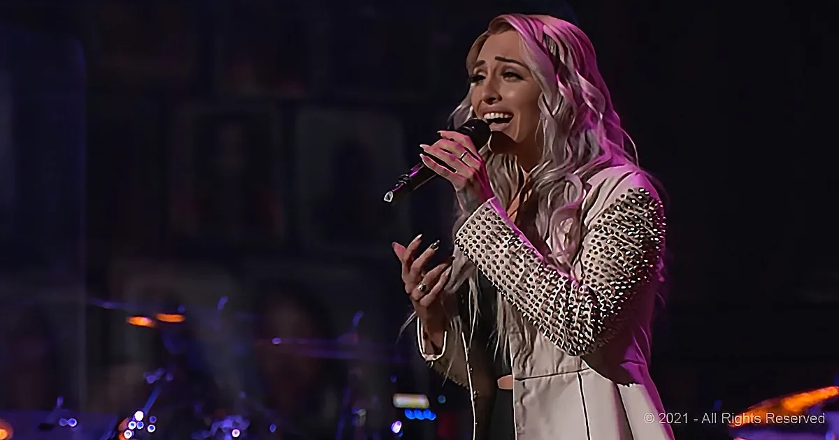 A woman with long, wavy hair sings passionately into a microphone, wearing a studded jacket and white pants.
