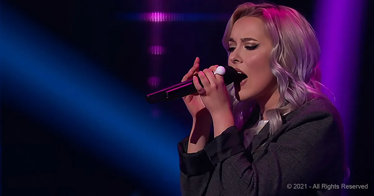 A woman with blonde hair sings into a microphone on stage under purple and blue lights.