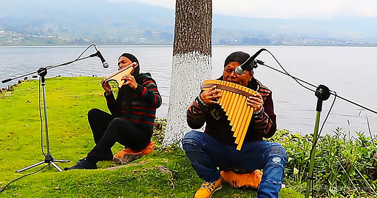 Two individuals play traditional instruments under a tree near a lake, with microphones set up.