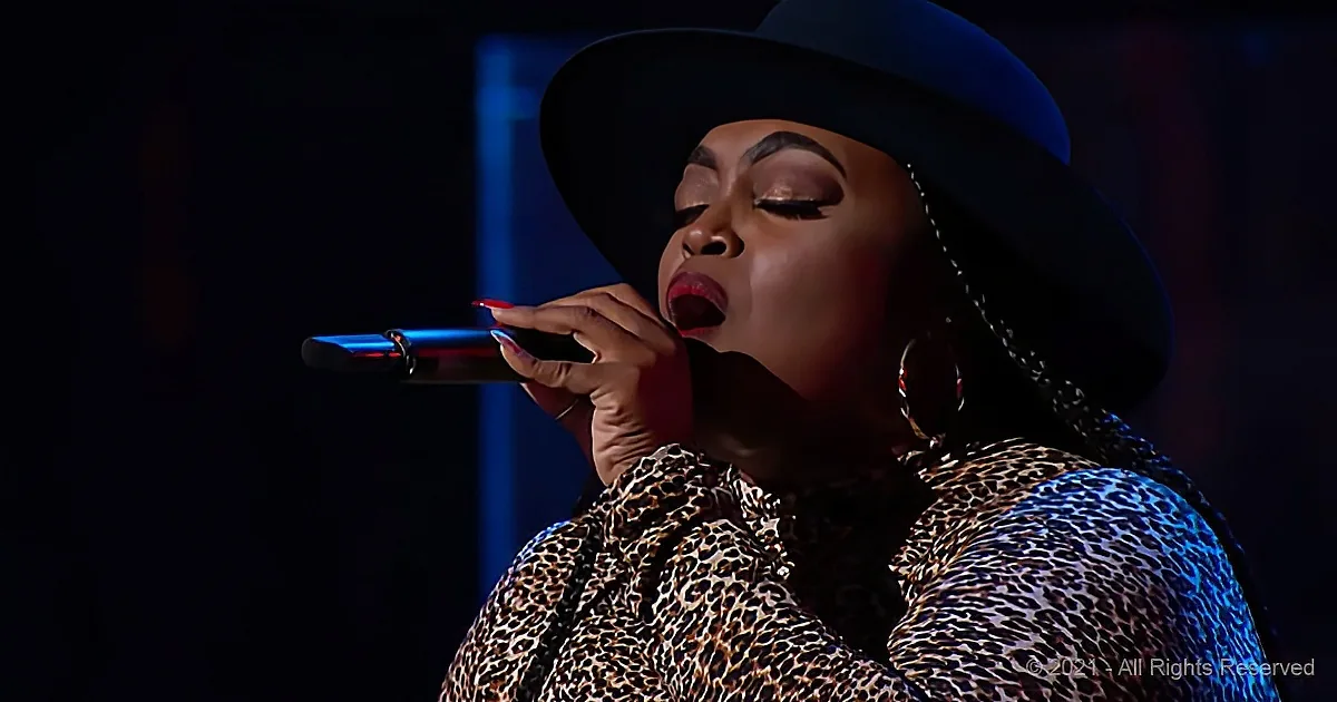 A woman in a leopard print top and black hat sings passionately into a microphone.