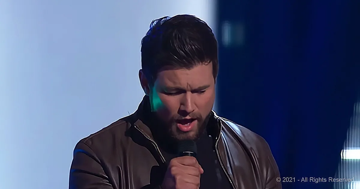 A man in a brown jacket sings into a microphone under blue stage lighting.