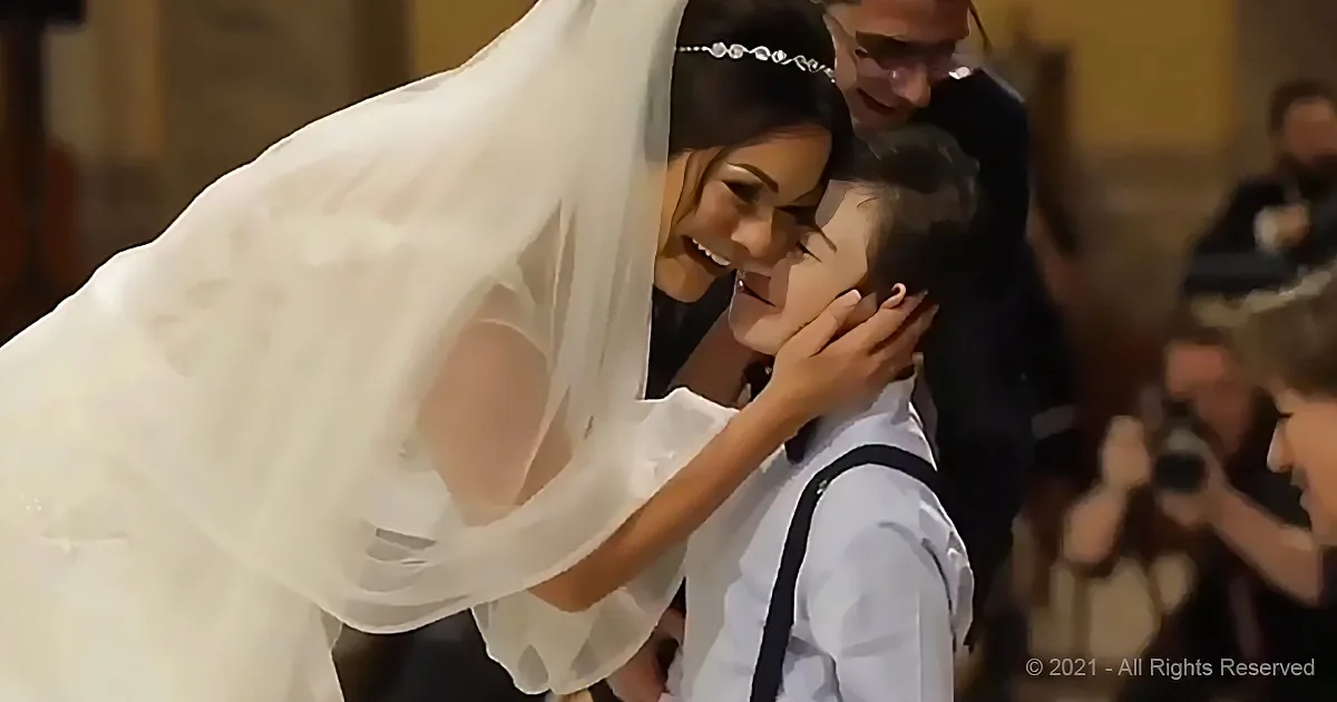 A bride in a white dress and veil is tenderly holding a young boy's face, smiling warmly.